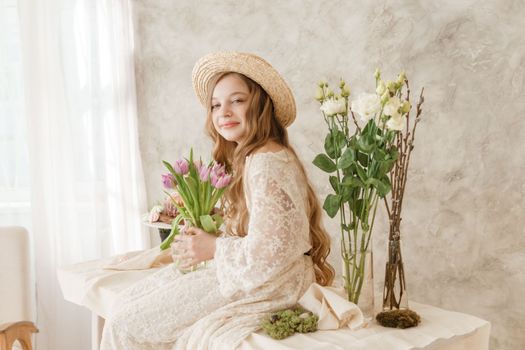 A girl with long hair in a dress and hat is sitting at the Easter table with cakes, spring flowers and quail eggs. Happy Easter celebration