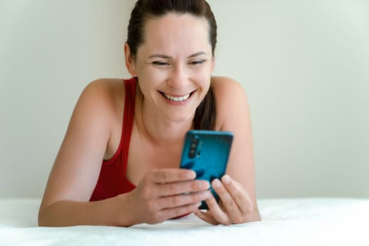 Girl with smartphone in her hands communicates via video communication with smile on her face lying on bed at home.