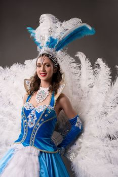 Positive young female in vintage blue dress with decorative shiny embellishments of crystals and diamonds with necklace and carnival headdress with feathers looking at camera