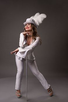 Full body of smiling young female in art deco suit with feathers on hat leaning on white cane standing against gray background looking up