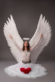 Young female sitting in studio on gray background wearing white dress angel wings and halo with red heart cushion raising hands