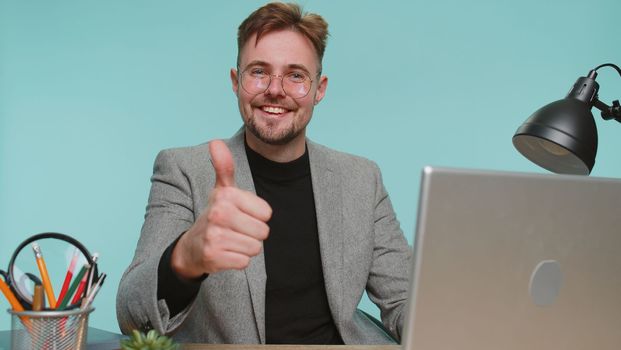 Like. Happy bearded business man sitting at office desk raises thumbs up agrees with something or gives positive reply recommends advertisement likes good. Young guy boy on blue studio background