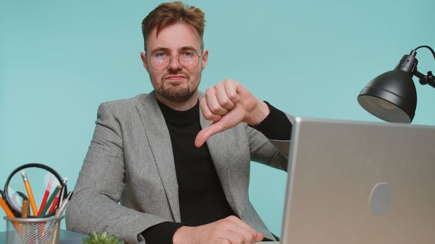 Dislike. Upset bearded business man sitting at office desk showing thumbs down sign gesture, expressing discontent, disapproval, dissatisfied, dislike, bad news. Young guy on blue studio background