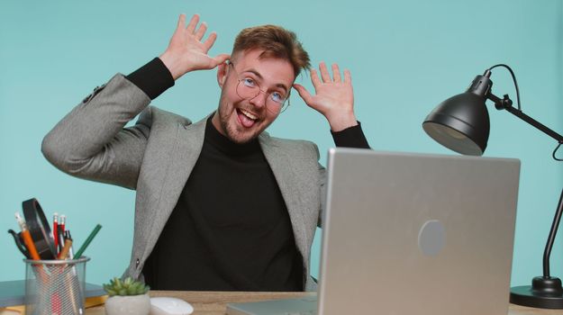 Cheerful funny bearded business man sitting at office desk showing tongue making faces at camera, fooling around, joking, aping with silly face, teasing. Young guy boy on blue studio background