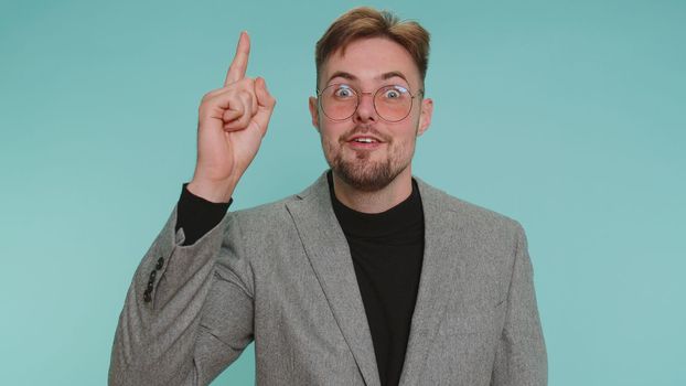 Eureka. Inspired business man in jacket pointing finger up with open mouth, showing Eureka gesture, solution, idea, inspiration, answer. Young adult guy isolated alone on blue studio wall background