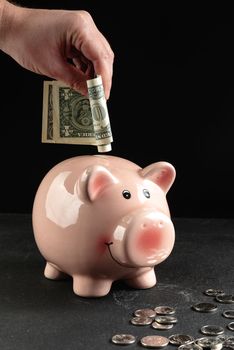 A man puts dollars in a piggy bank with a stack of coins next to it on a concrete table on a dark black background with copy space, using it as a business or financial savings concept