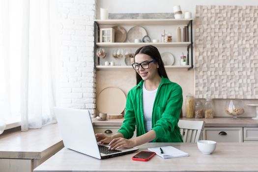 A young woman journalist, writer, blogger works remotely from home using a laptop online. Sitting in the kitchen at the table, making notes in a notebook.