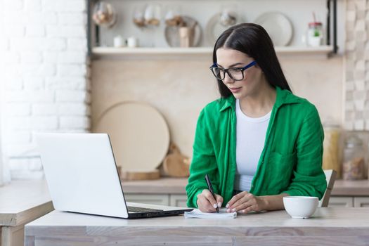 A young serious woman is having a remote job interview online. Sitting at home in the kitchen with a laptop. Speaks through a video call, introduces himself, passes the exam, writes in a notebook.