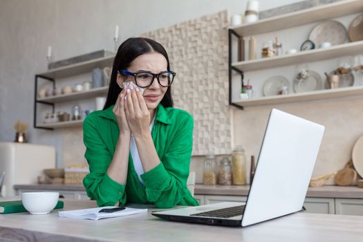 A young upset woman sits at home in the kitchen with a laptop, cries, wipes her tears with a napkin. Received bad news, watches a movie, talks on a video call.