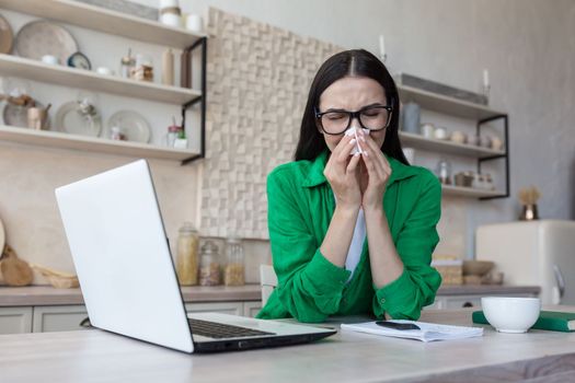 Tired young female freelancer student sitting at home in kitchen with laptop. Works. studies online. sick, feels bad, wipes the runny nose with a napkin.