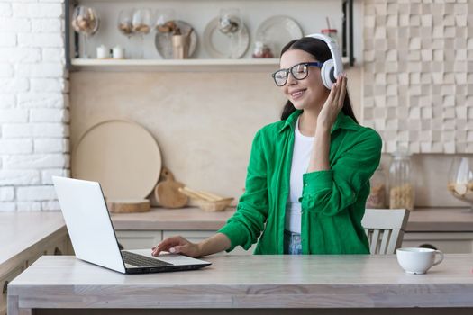 Young beautiful woman sitting in headphones at home at kitchen table and using laptop, Watch online webinar, movie, study, listens to music.