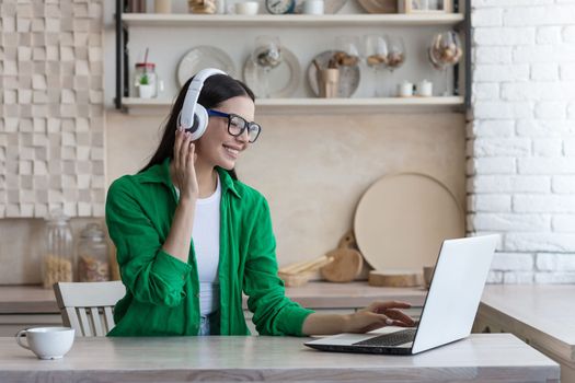 Young beautiful woman sitting in headphones at home at kitchen table and using laptop, Watch online webinar, movie, study.