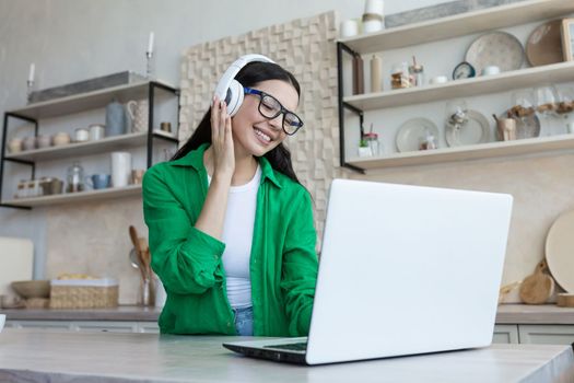 Young beautiful woman sitting in headphones at home at kitchen table and using laptop. Listens to music, enjoys, rests, smiles.