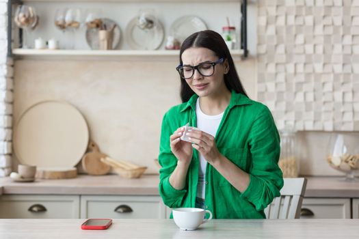 Sad young woman sits at home in the kitchen and cries, suffers. He wipes his nose and tears with a napkin. I received a message on the phone about divorce, bad news.