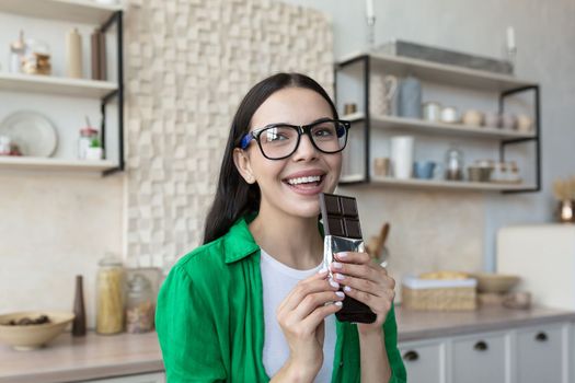 Portrait of a young beautiful woman holding and eat a bar of black chocolate in her hands. Standing at home in the kitchen, looking at the camera, smiling.