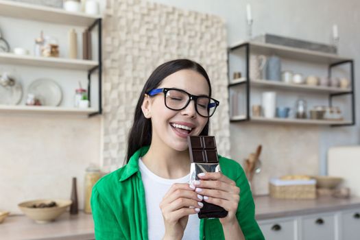 Portrait of a young beautiful woman eating a bar of dark chocolate. Standing at home in the kitchen, eyes closed, enjoying