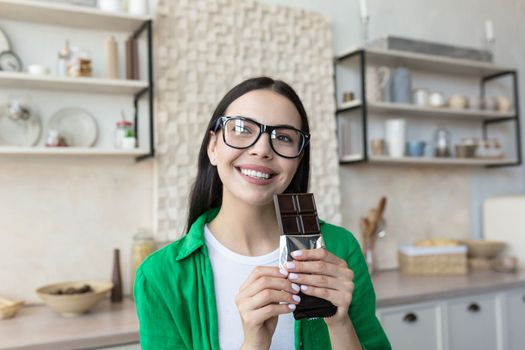 Close up photo. Portrait of a young beautiful woman holding a bar of black chocolate in her hands. Standing at home in the kitchen, looking at the camera, smiling.