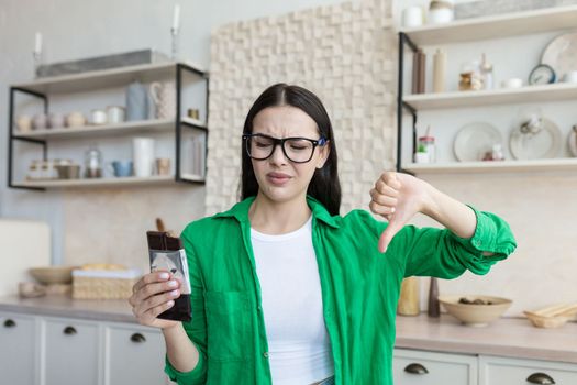 Dissatisfied young woman at home in the kitchen holding a bar of chocolate in her hand. With the other hand, he points a finger of dislike. Hates chocolate, sweets, supports a diet, counts calories.