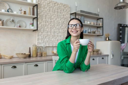 A young woman, businesswoman, freelancer, student drinks coffee and tea alone in the kitchen at home. Has a break. He holds a cup in his hands, rests, smiles, looks thoughtfully out the window.