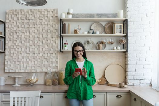 Happy and smiling young teenage girl standing at home in the kitchen and using the phone. Receives messages, chats with friends, boyfriend, chats news, blogs, social networks, online shopping.