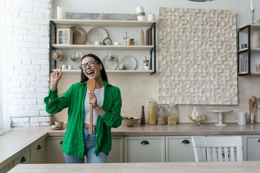 Joyful and happy young woman, housewife, student sings in the kitchen into a kitchen object as a microphone and dances. Is cooking.