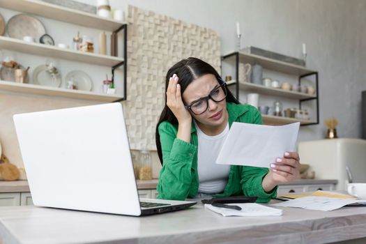 Shocked young woman working in kitchen at table with laptop. She holds a letter, a document in her hand, received bad news, an account, debt, divorce papers. He holds his head worriedly.