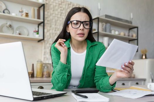 Worried young woman sitting in the kitchen at the table at home with a laptop. Holds a letter, a document in his hand, checks the account, debt, taxation.