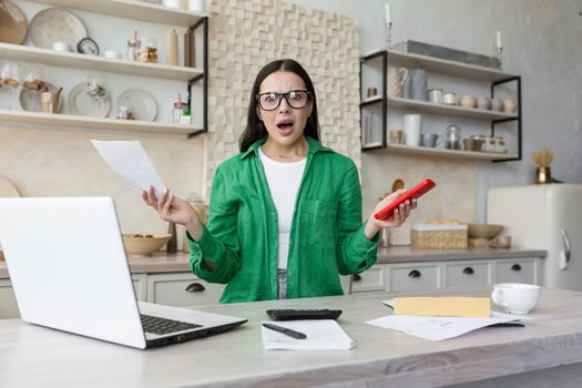 Portrait of worried and shocked woman sitting at home in kitchen with laptop and documents. Problems with accounts, credit, rent payment.