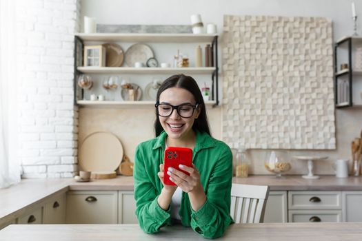 Happy and smiling young teenage girl sittiing at home in the kitchen and using the phone. Receives messages, chats with friends, boyfriend, chats news, blogs, social networks, online shopping.