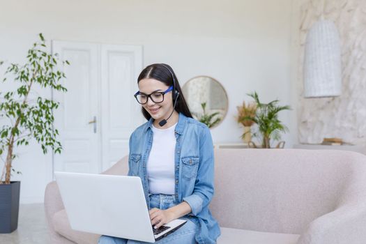 Beautiful young female student sitting on sofa at home with laptop and headset. He studies online, remotely.