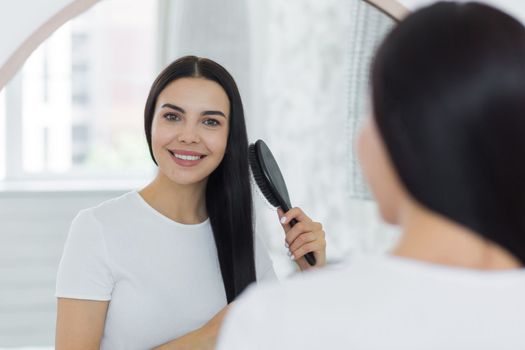 Close-up photo. A beautiful young woman stands and looks at herself in the mirror. She combs her hair with a brush, does her hair. She smiles, looking at the camera.