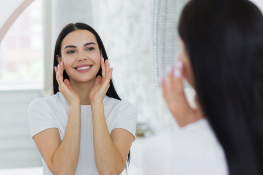 A beautiful young woman stands and looks at herself in the mirror. She touches her face with her hands, applies cream, cosmetics, does a massage, admires herself, smiles.