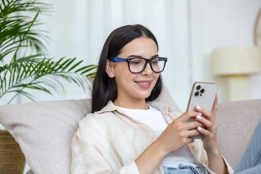 A young beautiful woman is lying on the sofa at home with a phone in her hands. Makes food orders for home, shopping in an online store.