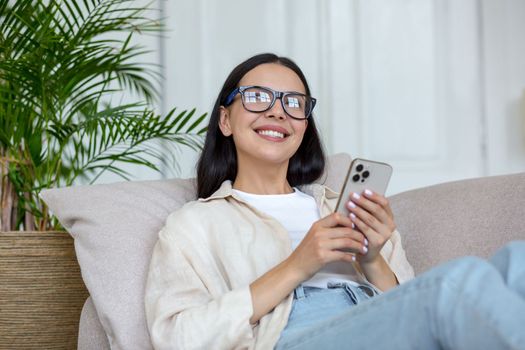 Happy young teenage girl lying on sofa at home and using phone. Receives messages, chats with friends, boyfriend, chat news, blogs, social networks, online shopping, looks at the camera, smiles