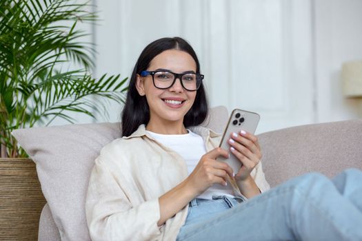 Young beautiful woman in glasses is lying on sofa and using mobile phone, looking at camera, smiling.