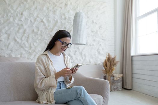 A young beautiful woman in glasses sits on a sofa by the window and uses a mobile phone, dials, texts, reads the news, smiles.