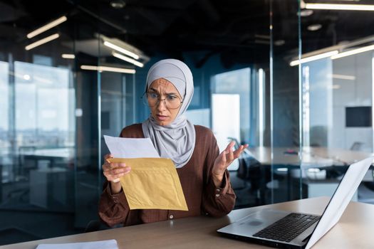 Upset business woman in hijab working inside office, woman received letter envelope mail with bad news, office worker upset by notification working at workplace with laptop.