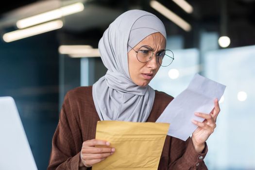 Upset business woman in hijab working inside office, woman received letter envelope mail with bad news, office worker upset by notification working at workplace with laptop.