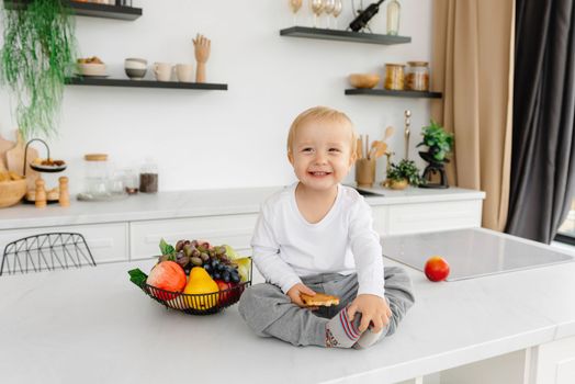 A happy child sitting in the kitchen smiling next to fruits and vegetables.