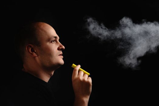 A young man smokes an electronic cigarette against a black background and blows smoke. E-cigarette safe or not, synthetic nicotine.