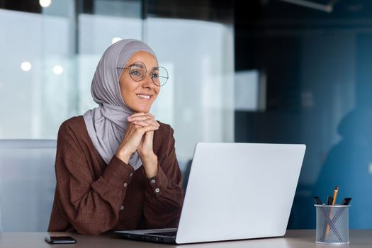 Smiling and dreamy businesswoman working inside office with laptop, woman in hijab and glasses office worker happy and satisfied with work sitting at desk.