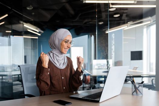 Successful businesswoman in hijab celebrating victory and successful achievement of results, woman looking at laptop screen and holding hands up in win and triumph gesture.