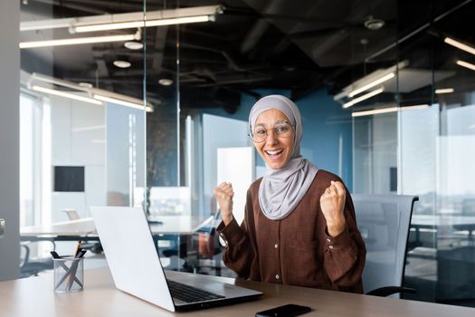 Portrait of successful businesswoman in hijab, muslim woman working inside office using laptop at work looking at camera and smiling happy, female worker celebrating victory good achievement results