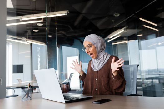 Successful businesswoman in hijab celebrating victory and successful achievement of results, woman looking at laptop screen and holding hands up in win and triumph gesture.