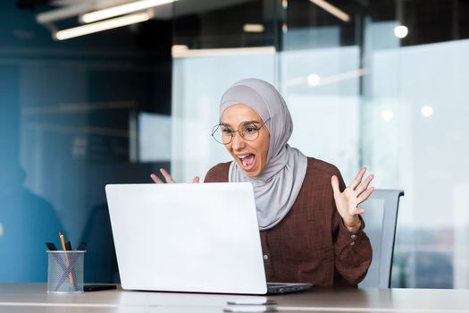 Successful businesswoman in hijab celebrating victory and successful achievement of results, woman looking at laptop screen and holding hands up in win and triumph gesture.