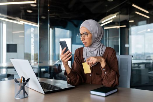 Upset and disappointed woman in hijab inside office trying to make purchase in online store, business woman holding smartphone and bank credit card, received money transfer error.