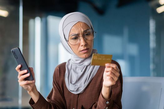 Upset and disappointed woman in hijab inside office trying to make purchase in online store, business woman holding smartphone and bank credit card, received money transfer error.