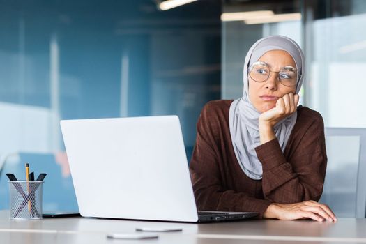 Serious bored businesswoman inside office, muslim woman in hijab thinking while sitting at workplace with laptop, woman at work thinking about decisions.