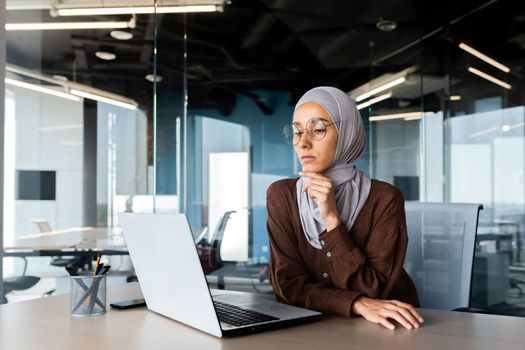 Serious bored businesswoman inside office, muslim woman in hijab thinking while sitting at workplace with laptop, woman at work thinking about decisions.