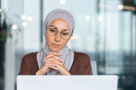 Serious bored businesswoman inside office, muslim woman in hijab thinking while sitting at workplace with laptop, woman at work thinking about decisions.